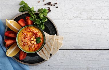 Geleneksel ev yapımı humus, zeytinyağı ve taze sebzeli kase. Tabakta limon ve pide dilimleri var. Faydalı doğal atıştırmalıklar. Ahşap beyaz tahtaların arka planı. Üst Manzara. Boşluğu kopyala