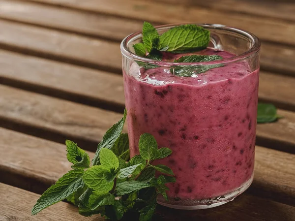 Blackcurrant smoothie with banana and mint in a glass cup. Smoothie decorated with mint leaves, a glass stands on a brown wooden table. Close-up.