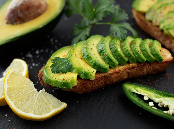 Avokado ile sandviç, siyah taşlı arka planda taze kesilmiş avokadolardan yapılan ev yapımı koyu bir ekmek. Yakın plan. Limon dilimleri, kişniş ve jalapeno ve arka planda yarım avacado. Hori.