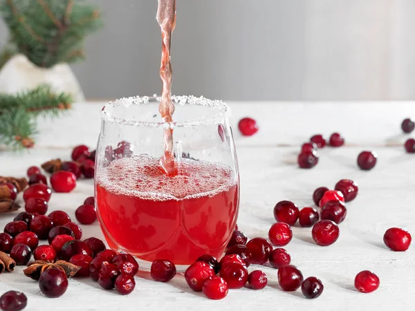 Verse cranberrydrank wordt in een glazen beker gegoten. Houten witte achtergrond. Een close-up. Horizontale oriëntatie. Kruisbessen en specerijen op tafel. — Stockfoto