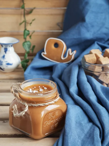 Kırsal bir ahşap masada cam bir kavanozda ev yapımı tuzlu karamel sosu. Yakın plan. Arka planda zencefilli kurabiye var. Seçici odaklanma, dikey yönelim