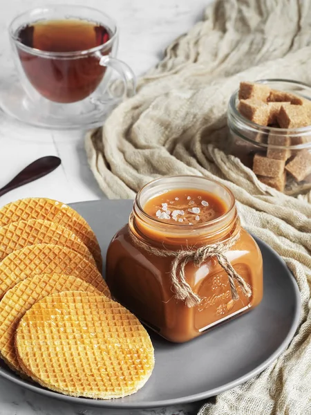 Homemade salted caramel sauce in a glass jar on a rustic wooden table. Close-up. Round caramel poured wafers in the foreground. Selective Focus, Vertical Orientation