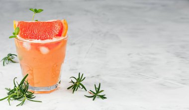 Cold lemonade of fresh grapefruit juice with alcohol or wihtout alcohole and ice cubes decorated a slice of grapefruit, twig mint and rosemary on the gray background. Copy space. Close up/