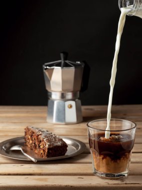 Süt bir bardak kahveye dökülür. Havuçlu kek tabağı. Kahve kahverengi ahşap bir arka planda yer almaktadır. Yakın çekim, dikey oryantasyon. Yüzeyin derinliği. Kahvaltı ya da atıştırmalık konsepti.