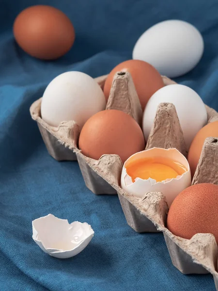 Chicken eggs in the cartom box. Chicken egg is half broken among other eggs, eggshell near. Close-up. Blue tablecloth background. — Stock Photo, Image