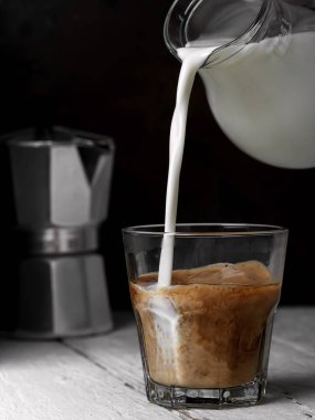 Beyaz ahşap zemin üzerinde espresso ve süt olan bir bardak. Süt bir bardak kahveye dökülür. Yakın çekim, dikey oryantasyon. Yüzeyin derinliği. Arka planda Macchinetta (kahve demliği).