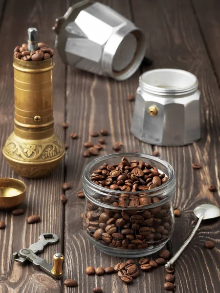 Granos de café tostados en un frasco de vidrio y granos de café dispersos en una mesa marrón de madera. Un molino de mano y una cafetera (la moka) en el fondo. Close Up, concepto de preparación de café tradicional . — Foto de Stock