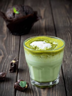 Süt köpüklü kibrit latte, tahta, siyah arka planda bir bardak. Yakınlarda çikolata parçaları var. Arka planda çikolatalı kek. Yakın çekim, yumuşak odaklanma.