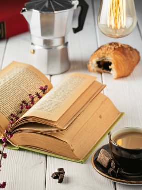 Taze kruvasan, espresso ve açık kitapla kahvaltı. Kitabın yanında bir buket çiçek var. Yakın çekim, alan derinliği.