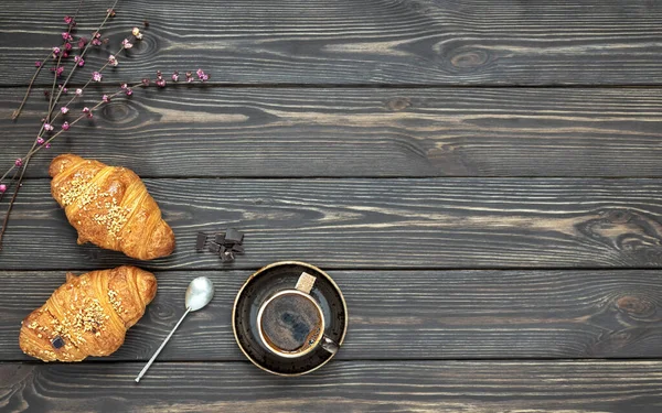 Kahvaltı sahnesi, taze kruvasanlar ve bir fincan espresso. Masada bir buket bahar çiçeği var. Koyu ahşap arka plan. Üst görünüm.