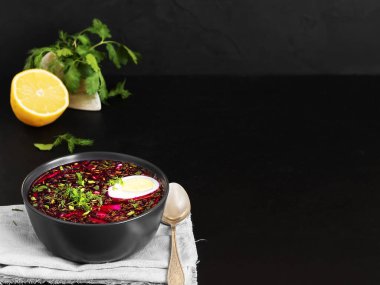 Yarım haşlanmış yumurtayla birlikte siyah seramik tabakta pancar çorbası. Gri peçetede bir kase, kaşığın yanında, yarım limon ve kişniş. Uzayı kopyala Siyah arka plan. Sağlıklı, doğal yiyecekler. Yakın plan..