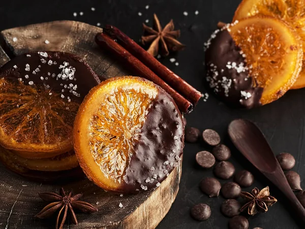 Christmas sweets, candied fruits in chocolate. Caramelized orange slices on a wooden stand, black stone background. Near spices and chocolate. Homemade Christmas dessert. Close-up.
