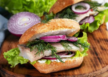 İskandinav mutfağı, deniz ürünleri burger, taze kızarmış çörek, ringa filetosu, soğan ve marul. Tabak, ahşap bir tahtanın üzerinde servis edildi. Yakın çekim, alan derinliği.