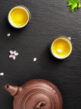 Yeşil Japon çayı seramik çaydanlıkta ve siyah taş bir masada. Masada sakura çiçekleri ve taç yaprakları var. Üst manzara, çay seremonisi, minimalizm.