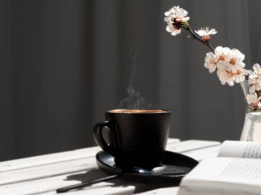 Açık bir kitap ve çiçek açan sakura dalının yanında, ahşap bir masanın üzerinde aromatik espresso ile bir fincan kahve. Yakın plan. Sıcak kahvenin üstünde buhar yükselir..