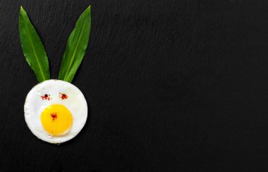 Eğlenceli bir Paskalya kahvaltısı fikri kızarmış yumurtadan yapılmış bir Paskalya tavşanı, yabani sarımsak yapraklarından yapılmış tavşan kulaklarıdır. Siyah taş arkaplan, üst görünüm, metin için yer, en düşük biçim