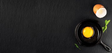 Çiğ tavuk yumurtası, dökme demir ya da çelik bir tavada, koyu taştan bir arka planda kırılmış. Kabuğunun ve roka yapraklarının yanında parlak bir yumurta sarısının üst görüntüsü, tarif için alanı kopyalamak. Kahvaltı ya da öğle yemeği konsepti