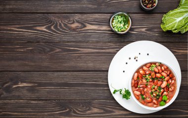 Beyaz tabakta fesleğen yapraklarıyla süslenmiş haşlanmış kırmızı fasulye. Baharat ve taze salatanın yanında sağlıklı vejetaryen yemeği. Koyu renkli ahşap masa üzerinde düz bir görünüm, metin için kopya alanı ile yatay görünüm