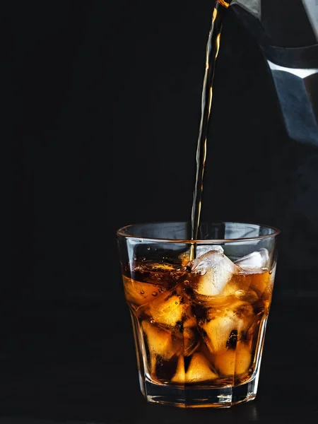 Iced coffee in a glass with ice and sugar syrup. Coffee is poured from a coffee pot into a glass. A refreshing and invigorating drink in summer in hot weather. Close-up, dark wood background.