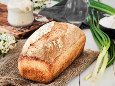 Çuval bezinde taze pişmiş ekmek, ekşi ekmek ve beyaz ahşap bir masada bir sürahi suyla un. Hobiler, evde buğday ekmeği pişirmek. Sağlıklı gıda konsepti, geleneksel zanaat ekmeği. Yakın çekim.
