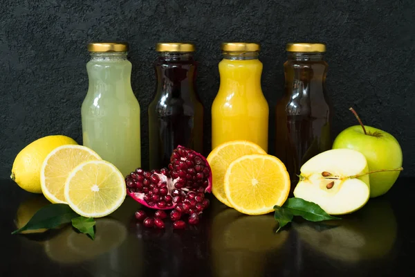 Four types of juice (pomegranate, apple, lemon, orange juice) in glass bottles on a black wooden background.