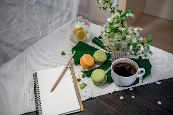 Crushed pastel, orange and green macaroons on a wood background with whole one in white plate with jug with flowers on the back