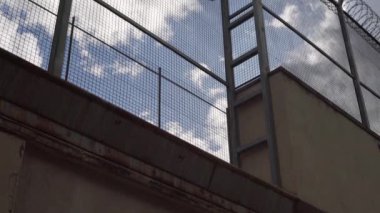 Mavi gökyüzüne dikenli tellerle çevrili yüksek hapishane çitleri. Kamera yavaşça yükseliyor.