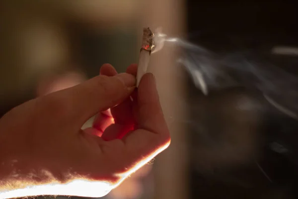 A hand holding a burning marijuana joint with plumes of smoke rising from it.