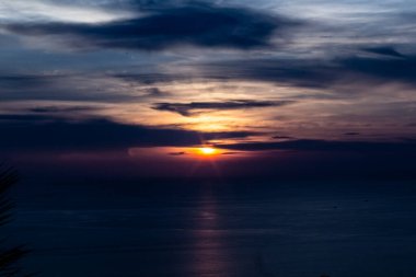 Phuket Tayland 'da Karon' un tepesinden görülen karanlık ve canlı bir gün batımı. Derin turuncu bir güneş okyanusun üzerinde ağır bulutların ardında batıyor..