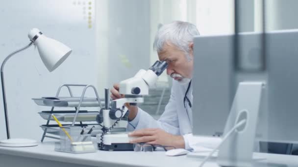 シニア男性医師の健康を調査する顕微鏡を使って関連する事項について。デスクトップ コンピューターで近くのアシスタント作品. — ストック動画