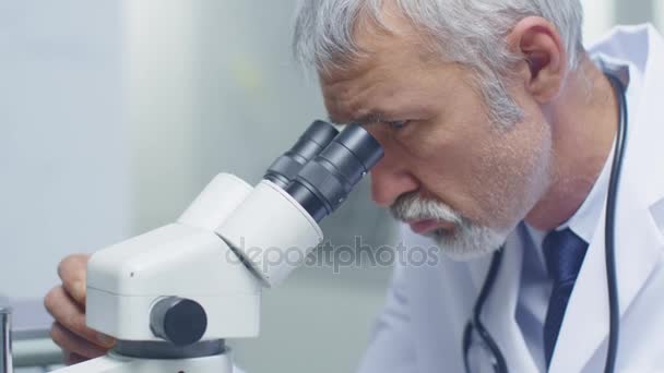 Senior muž lékař provádějící zdravotní problém šetření přes mikroskop. Jeho asistent pracující na počítači nedaleké. — Stock video