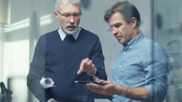 Två seniora ingenjörer diskutera tekniska detaljer över tablett dator. Båda männen ser respektabel och professionella. Office är ljusa och moderna. — Stockvideo