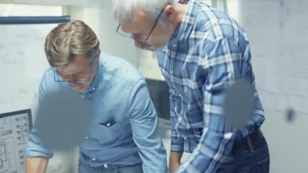 Dos ingenieros de arquitectura senior examinan planos mientras están de pie sobre la mesa de trabajo. Ellas tienen discusiones sobre el tema de varios asuntos técnicos concernientes al proyecto pendiente . — Vídeos de Stock