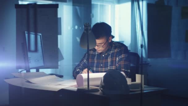 Jeune ingénieur en architecture compétent travaillant tard dans son bureau. Le bureau est sombre seulement sa lumière de table est allumée. Divers plans et plans reposent sur la table. Il écrit ses calculs dans son bloc-notes . — Video