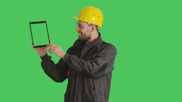 中期拍摄的英俊的工人戴着安全帽制作刷触摸手势与另一个平板电脑用一只手按住。平板电脑和背景是绿色屏幕. — 图库视频影像