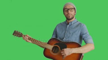 Orta bir kadeh bir genç yetenekli müzisyen şapka ve gözlük gitar çalmak. Yeşil ekran arka plan olduğunu.