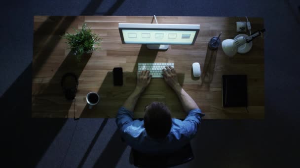 Bovenaanzicht van jonge creatieve ontwerper werken op zijn bureaubladcomputer bij nacht. Zijn tafel wordt verlicht door koude blauw licht van buiten. — Stockvideo