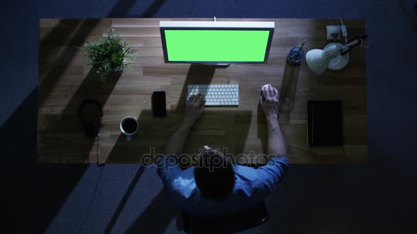 Visão superior do programador masculino que trabalha em seu computador desktop com tela verde da falsificação na noite. Sua mesa é iluminada pela luz azul fria de fora . — Vídeo de Stock