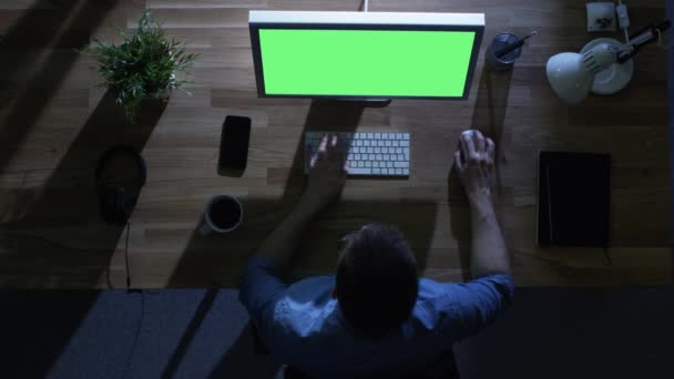 顶视图的男性程序员在工作与模拟绿色他桌面电脑屏幕在晚上。他喝咖啡。他的桌子被照亮冷蓝色光从外面. — 图库视频影像