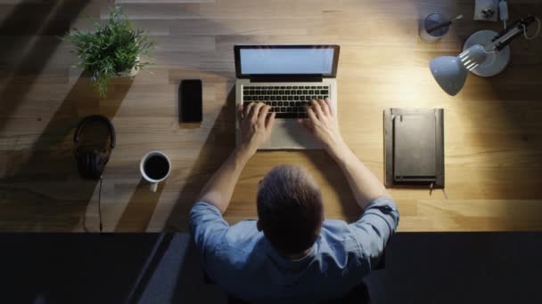 Vista dall'alto del giovane designer creativo che lavora sul suo computer portatile di notte. Lui beve del caffè. Lampada da tavolo accesa . — Video Stock