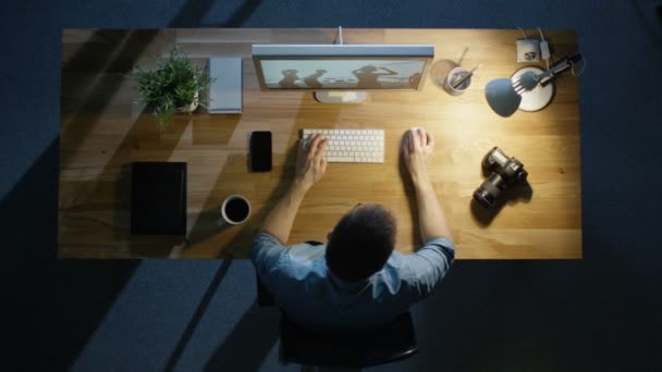 Vue du dessus d'un jeune photographe traitant des photographies sur son ordinateur de bureau tard dans la nuit. Lampe de table illumine son bureau. Appareil photo, disque dur externe et ordinateur portable couché à côté de lui . — Video