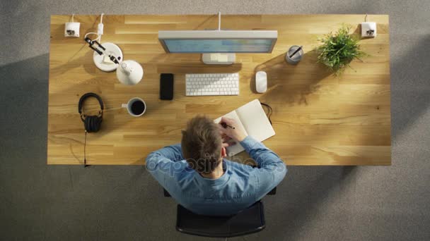 Ansicht von oben des kreativen Arbeiters, der während der Arbeit an seinem Desktop-Computer das arbeitsbezogene Notizbuch füllt. auch auf der Tischplatte befinden sich Smartphone, Kaffeetasse, Kopfhörer, Pflanze. — Stockvideo