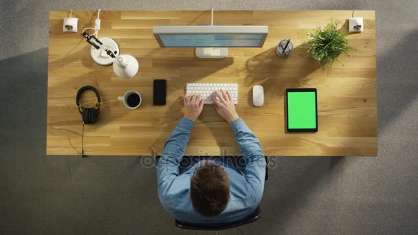 Vista dall'alto di un ingegnere del software che lavora al suo computer desktop seduto nel suo ufficio creativo. Sdraiato sul tavolo: Smartphone, Tablet Computer, Cuffie . — Video Stock