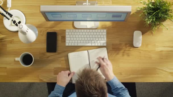 Vista dall'alto dell'ingegnere del software che annota l'elenco dei lavori correlati nel suo taccuino mentre lavora al suo computer desktop . — Video Stock