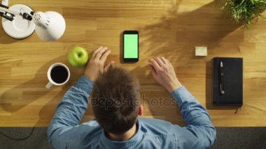 Masasında otururken genç yaratıcı adam dokunarak onun yeşil ekran smartphone cep telefonu ile Üstten Görünüm.