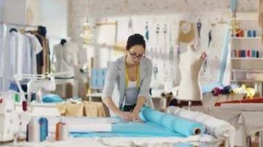 Güzel kadın moda tasarımcısı bir kumaş parçası üzerinde çizer. O bir ışık renkli stüdyoda çalışıyor dolu çeşitli giysiler, kumaşlar ve çizimler.