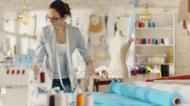 Güzel kadın moda tasarımcısı düzeltiyor rulo, mavi kumaşlar ve bırakır dışarı şablonları üstünde o. O bir ışık renkli stüdyoda çalışıyor tam çeşitli giysiler, kumaşlar ve duvardaki çizimler.