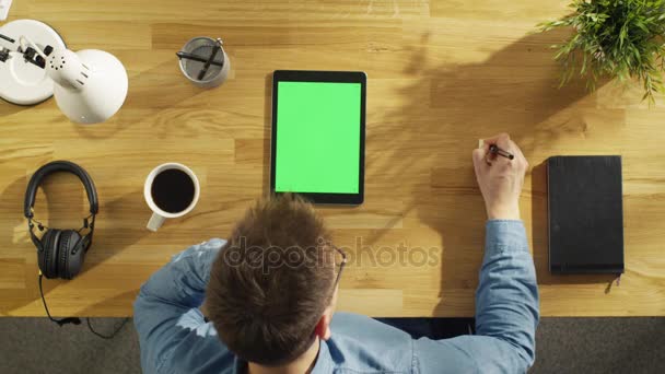 Ovanifrån av unga kreativa Man som arbetar på sin handdator som sitter vid sitt skrivbord. Hörlurar, Notebook, växt och kaffekopp är på bord. — Stockvideo