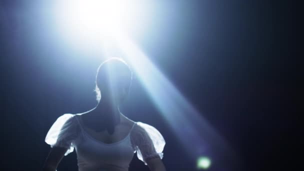 Mid Shot van een mooie jonge Ballerina sierlijk in de schijnwerper, duisternis om haar heen dansen. Ze is dragen witte Tutu jurk, dat in het licht schittert. In Slow Motion. — Stockvideo