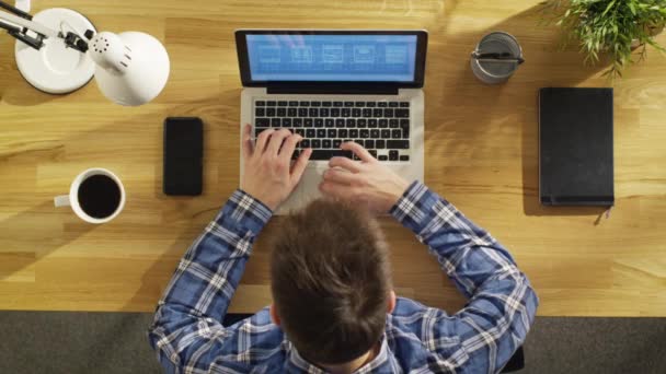 Vista superior de un joven creativo que trabaja en su computadora portátil mientras está sentado en su escritorio de madera. También en la mesa: Taza de café, Smartphone, Cuaderno, Lámpara, Planta . — Vídeos de Stock